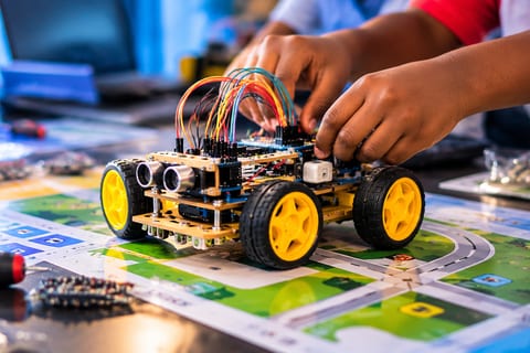 Illustration of hands connecting wires on a wheeled robot at a workshop table, with tools and electronic parts around it.